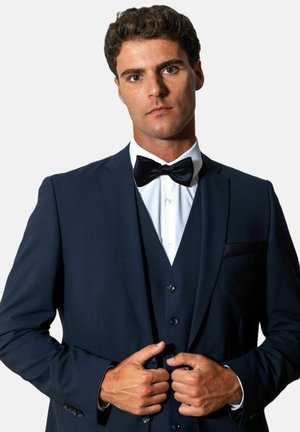 Homme boutonnant une veste de costume bleu marine, portant une chemise blanche, un gilet bleu marine et un nœud papillon noir, sur un fond clair uni.