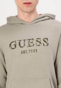 Grüne Kapuzenjacke mit auffälligem "GUESS"-Logo und "EST. 1981" in texturiertem Stickereidruck. Weicher Stoff im klassischen Schnitt und mit Kängurutasche.