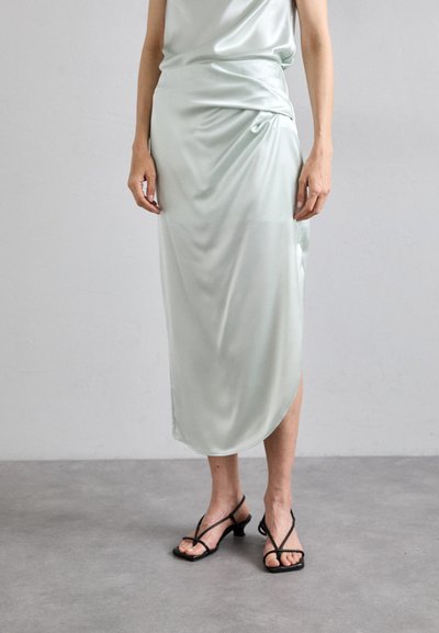 Mintgrünes Satin-Kleid mit drapiertem Design, knielang, mit einem seitlichen Schlitz. Kombiniert mit schwarzen, riemchen Sandalen mit hohen Absätzen.