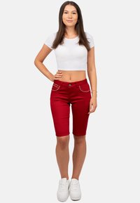 Pantaloni bermuda in denim rosso con cuciture a contrasto e due tasche frontali, abbinati a un crop top bianco aderente e scarpe bianche.