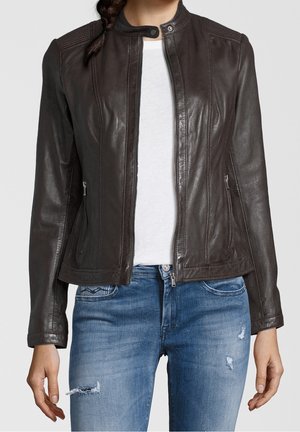 Frau trägt eine dunkelbraune Lederjacke über einem weißen Hemd mit blauen, abgenutzten Jeans und steht vor einem schlichten Hintergrund.