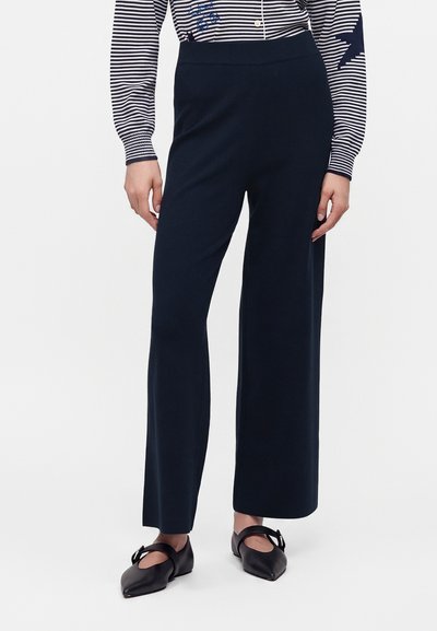 Femme portant un pantalon noir à jambes larges, des ballerines pointues noires avec une bride, et une chemise rayée à manches longues avec un motif étoile sur la manche.