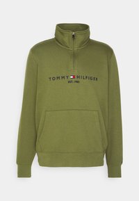 Sweatshirt vert olive à fermeture éclair à quart, avec poche avant, texte "Tommy Hilfiger EST. 1985" et logo brodés sur la poitrine.