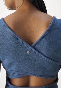 Femme aux cheveux tressés portant un haut bleu texturé à enroulement avec un dos ouvert et un petit bouton métallique rond.