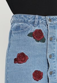 Jupe en denim de couleur bleu clair, ornée de roses bourgogne brodées et de feuilles vertes, fermeture à boutons à l'avant et deux poches avant.