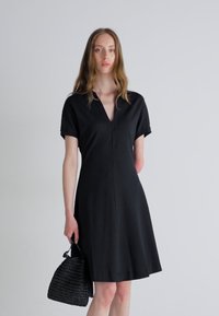 Robe noire à manches courtes avec un décolleté en V, une silhouette évasée et un tissu lisse. Accompagnée d'un sac noir texturé.