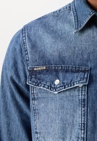 Gros plan sur la poche d'une veste en denim bleu avec rabat à bouton et petite étiquette métallique de la marque "Antony Morato".