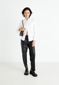 Camisa blanca de botón con un bolsillo en el pecho, combinada con pantalones de cuero negros y zapatos oscuros. El modelo sostiene un bolso cruzado marrón.