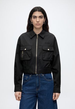 Femme aux longs cheveux foncés portant une veste noire à fermeture éclair avec poches à rabat à l'avant et un jean bleu taille haute, debout devant un fond uni.