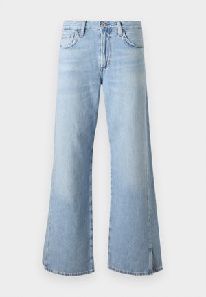 Zwart persoon staat met handen in de zakken, draagt een wit T-shirt, lichtblauwe wijde jeans en marineblauwe sneakers tegen een effen achtergrond.