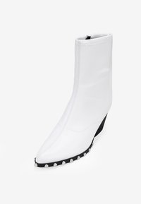 Bottine blanche en matériau synthétique lisse, dotée d'un bout pointu, d'un talon compensé et d'un accent noir avec des clous argentés le long de la semelle.