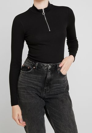 Personne portant un haut noir à manches longues avec une fermeture éclair argentée sur le devant et un jean noir délavé taille haute, main dans la poche.