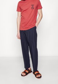 Camiseta de algodón roja con estampado negro, combinada con pantalones de pierna ancha azul marino y sandalias negras con correas naranjas y amarillas.