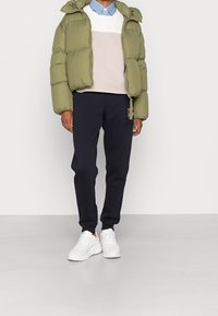 Osoba ubrana w oliwkową kurtkę puchową, beżowy i biały sweter, granatowe dresy z logo na boku oraz białe sneakersy, stojąca na szarym podłożu.