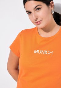 Oranges Baumwoll-T-Shirt mit kurzen Ärmeln und Rundhalsausschnitt. Mit weißem, gesticktem Text "MUNICH" auf der Brust. Glatte Textur.