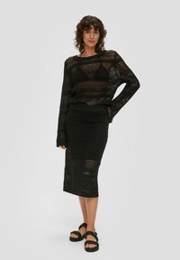 Czarny, dziergany sweter z siateczki z długimi rękawami i szerokim dekoltem, w połączeniu z dopasowaną czarną, wzorzystą spódnicą midi oraz masywnymi sandałami.