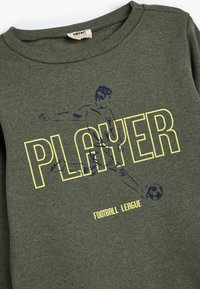 Sudadera de algodón verde con cuello de canalé, que presenta el texto amarillo "JUGADOR" y un gráfico de un futbolista. Textura suave.