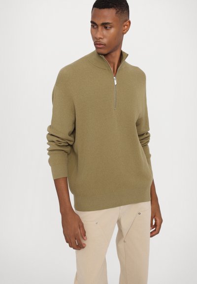 Olive green zippered sweater with ribbed texture, long sleeves, and relaxed fit. Paired with light beige trousers featuring snap details.