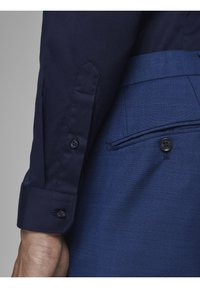 Námořnicky modrá košile s rukávem a knoflíkovým manžetou, spárovaná s modrými texturovanými kalhotami s přední kapsou a černým knoflíkovým detailem.