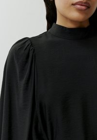 Zwarte blouse met een hoge halslijn, poffende mouwen en zachte, gestructureerde stof. Het ontwerp benadrukt drapering en vloeiende lijnen.