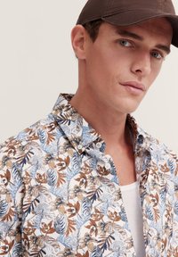 Blumenhemd mit Knopfleiste, das eine weiße Grundfarbe mit braunen und blauen Blattmustern zeigt, aus weichem Stoff und mit Kentkragen. Begleitet von einer braunen Mütze.