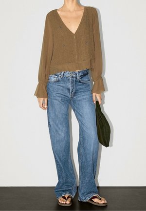 Femme portant une blouse marron à manches longues avec des poignets à volants, un jean bleu à jambes larges, des tongs, et tenant un sac à main vert foncé.