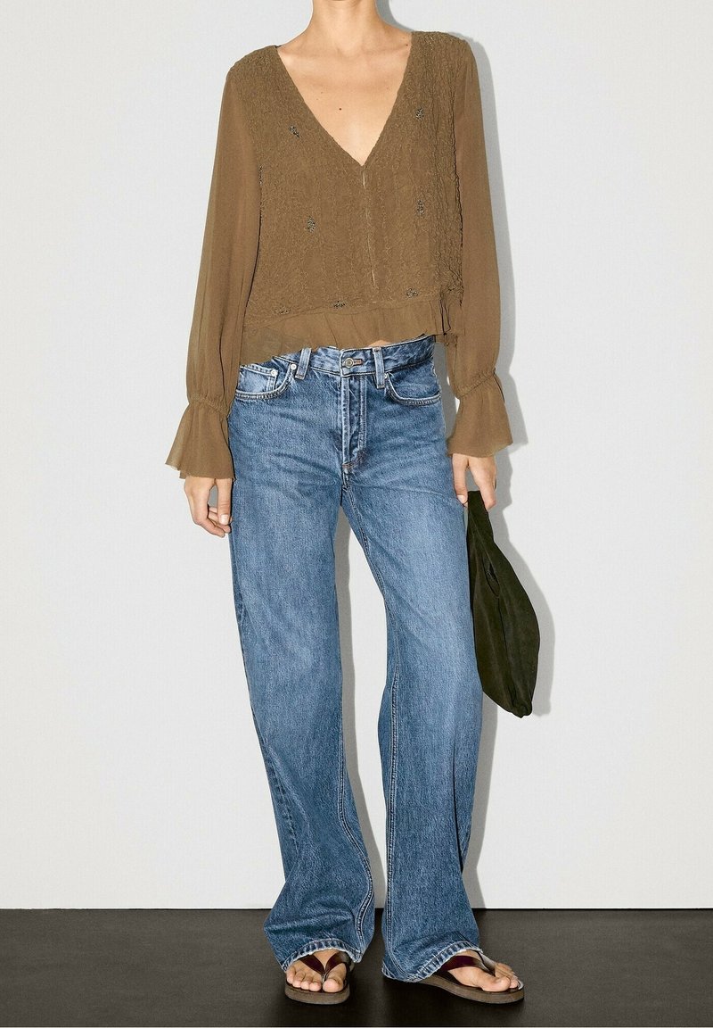 Femme portant une blouse marron à manches longues avec des poignets à volants, un jean bleu à jambes larges, des tongs, et tenant un sac à main vert foncé.