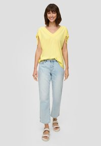 Gelbes V-Ausschnitt-Oberteil mit geknoteten Ärmeln, kombiniert mit hellblauen geraden Jeans und beigen Sandalen mit geflochtenen Riemen.