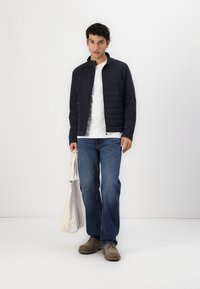 Giacca scura navy da uomo con parte anteriore trapuntata, T-shirt bianca, jeans blu, scarpe grigie e borsa a tracolla beige, in piedi contro uno sfondo neutro.