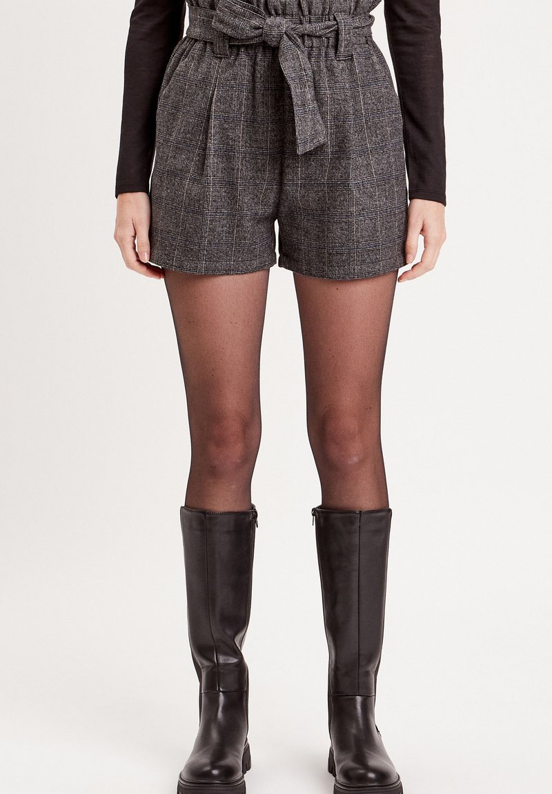 Shorts à carreaux gris avec taille haute et ceinture nouée, associés à des collants noirs transparents et des bottes en cuir noir jusqu'aux genoux.