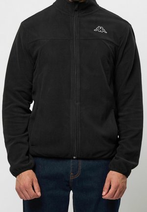 Mand iført en sort fleecejakke med lynlås og hvidt logo på brystet og blå jeans mod en ensfarvet baggrund.