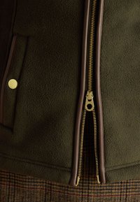 Dark green fleece jacket with gold zipper partially open, brown leather trim, and a buttoned pocket above checked fabric background.