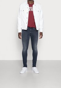 Chaqueta de mezclilla blanca sobre una camiseta burdeos con el estampado "HARVARD", combinada con jeans ajustados oscuros y zapatillas blancas, de pie sobre un fondo claro.