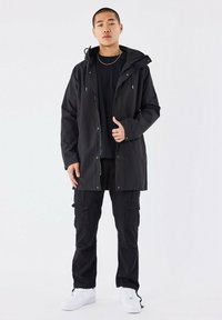 Giacca impermeabile nera con cappuccio regolabile, bottoni frontali e tasche; indossata sopra una maglietta nera e abbinata a pantaloni cargo neri e sneakers bianche.