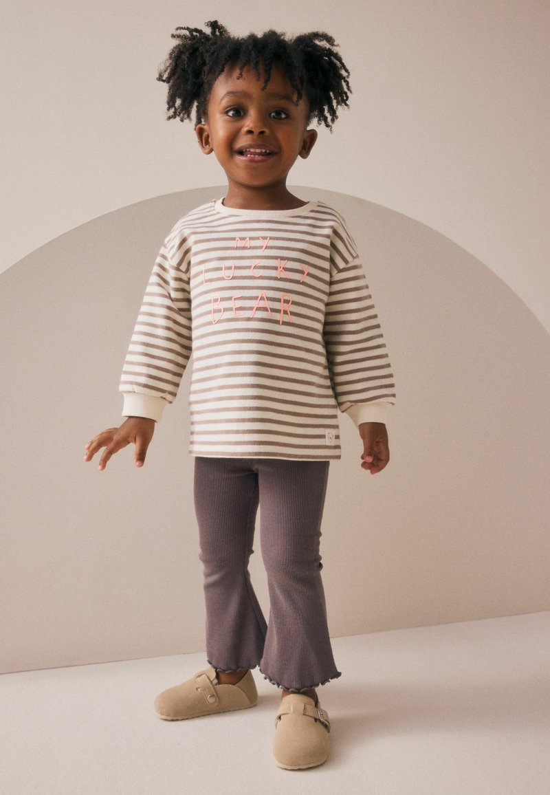 Maglietta a maniche lunghe a righe in crema e taupe con il testo "IL MIO ORSO FORTUNATO", abbinata a leggings a coste viola a zampa d'elefante e scarpe beige slip-on.