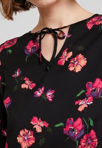 Schwarze Bluse mit floralem Muster, die einen V-Ausschnitt mit Bindeband aufweist und mit lebhaften pinken und lila Blumen auf einem glatten Stoff verziert ist.