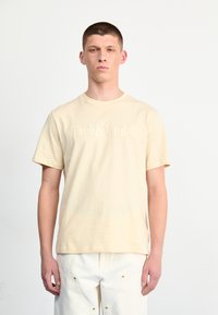 Beige katoenen t-shirt met korte mouwen en een ronde hals, met een reliëf "Timberland" logo op de voorkant. Draagt men met witte broeken.