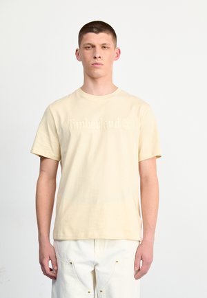 Beige cotton t-shirt with short sleeves and a crew neck, featuring embossed "Timberland" logo on the front. Worn with white pants.