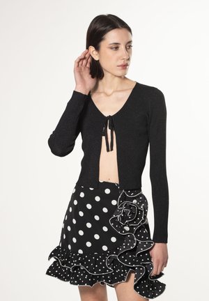 Sort strikket cropped cardigan med lange ærmer og bindefor, parret med en polkaprikket nederdel med flæser, der har sort og hvide mønstre.