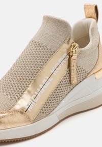 Sneaker con tomaia in maglia di beige chiaro con accenti dorati, branding bianco e una finitura testurizzata. Tallone imbottito e suola in gomma.