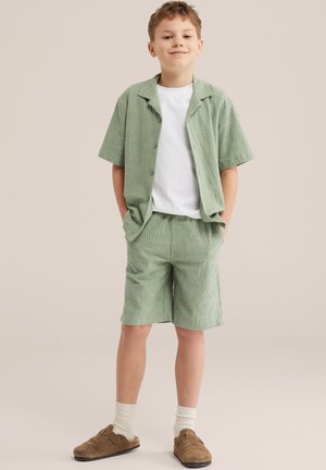 Garçon debout avec les mains dans les poches, portant une chemise à manches courtes texturée vert clair et un short assorti, un t-shirt blanc, des chaussettes blanches et des chaussures marron à enfiler.