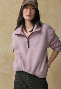 Svijetlopurple pulover od flisa s visokim ovratnikom i pola patentnog zatvarača, uparen s tamnim hlačama s uzorkom. Ima meku, strukturiranu tkaninu.