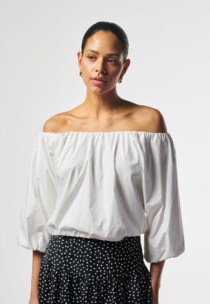 Vrouw met donker haar, gekleed in een witte off-shoulder blouse en een zwarte rok met witte stippen, staand tegen een effen lichte achtergrond.