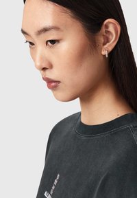 Ung kvinde med glat mørkt hår, iført en mørkegrå sweatshirt med rund hals og en lille sølvrektangelformet ørering på venstre øre.