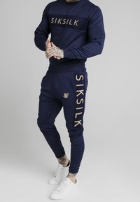 Man som bär en marinblå SikSilk-brandad träningsdräkt med vita sneakers, står mot en enkel ljus bakgrund.