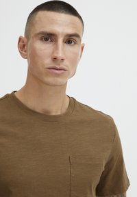 Brun kortermet T-skjorte laget av bomull, med rund hals og en liten brystlomme. Stoffet har en myk tekstur.