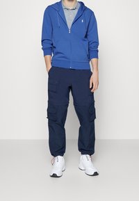 Blå zip-up huvtröja med kängurufickor, grå pikétröja, marinblå cargobyxor med flera fickor, och vita träningsskor.