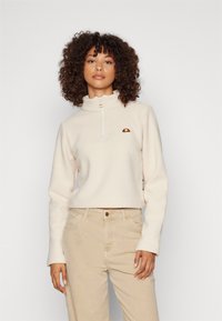 Kerman fleece puolipaita korkealla kauluksella, jossa on brodeerattu logo rinnassa ja ribbadut hihansuut, yhdistettynä vaaleisiin housuihin.