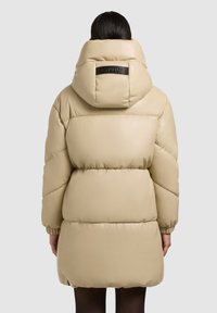 Béžová puffa bunda s vysokým golierom a kapucňou, s čiernym koženým logom na chrbte. Hladká, prešívaná textúra.