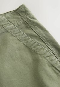 Gros plan d'un tissu vert montrant des coutures détaillées et des lignes de couture sur un vêtement, probablement un pantalon ou un short.